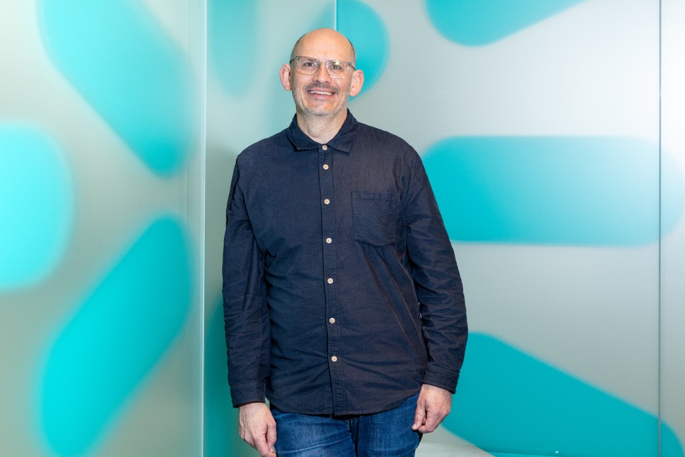 Smiling bald man wearing glasses, a dark button-up shirt, and jeans standing against a wall with turquoise and white abstract shapes.