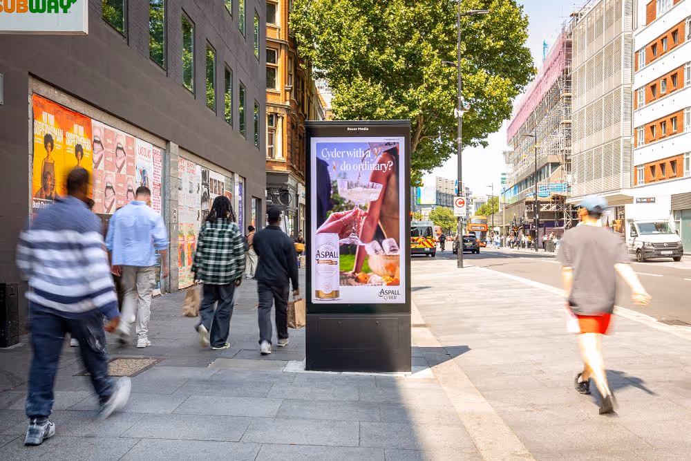Busy city street with pedestrians walking past a digital billboard displaying an Aspall Cider advertisement.