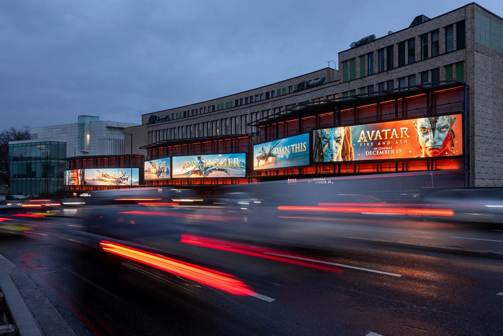 Building with illuminated billboards advertising the movie Avatar: Fire and Ash along a busy street with blurred moving cars at dusk.