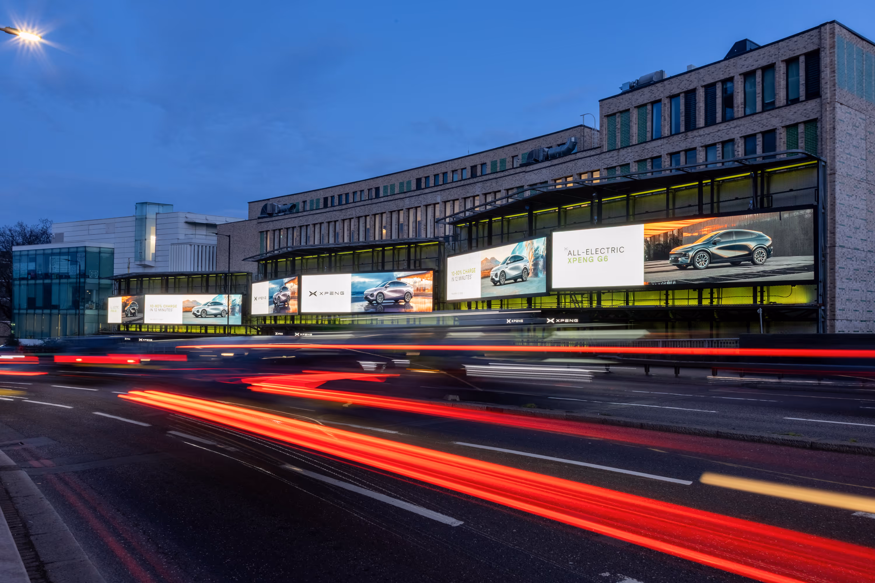 Evening city street with light trails of passing cars in front of a building featuring illuminated advertising panels for the all-electric XPENG G6 car.