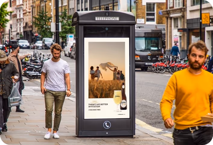 Outdoor digital phone booth display featuring an Aspall Cyder ad with people waving a cloth in a wheat field at sunset and a bottle with a glass of cyder.