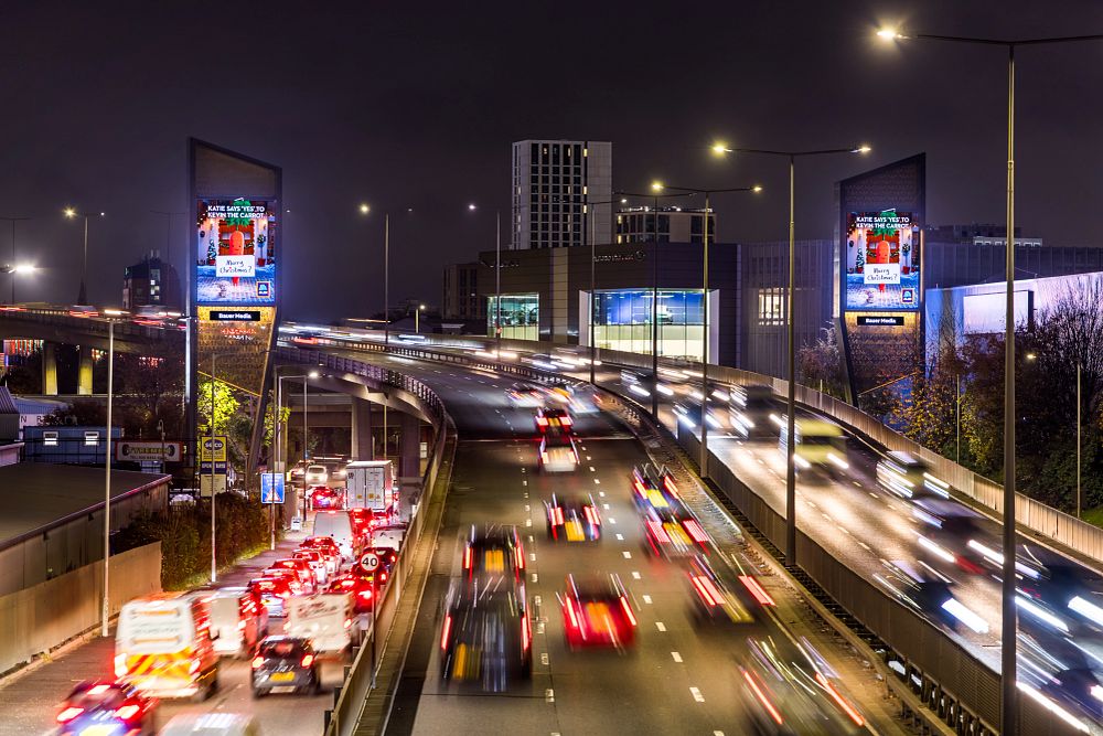 Nighttime highway scene with heavy traffic on the left lanes and fast-moving vehicles on the right lanes, flanked by illuminated digital billboards and streetlights.