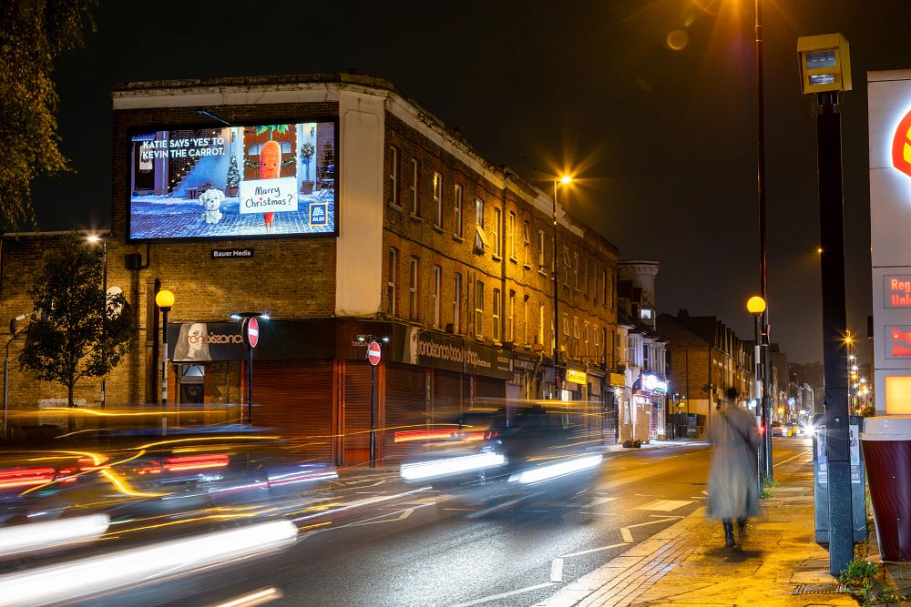 Night street scene with blurred car lights passing a brick building featuring a brightly lit billboard advertising Aldi with a Christmas theme and a person walking on the sidewalk.