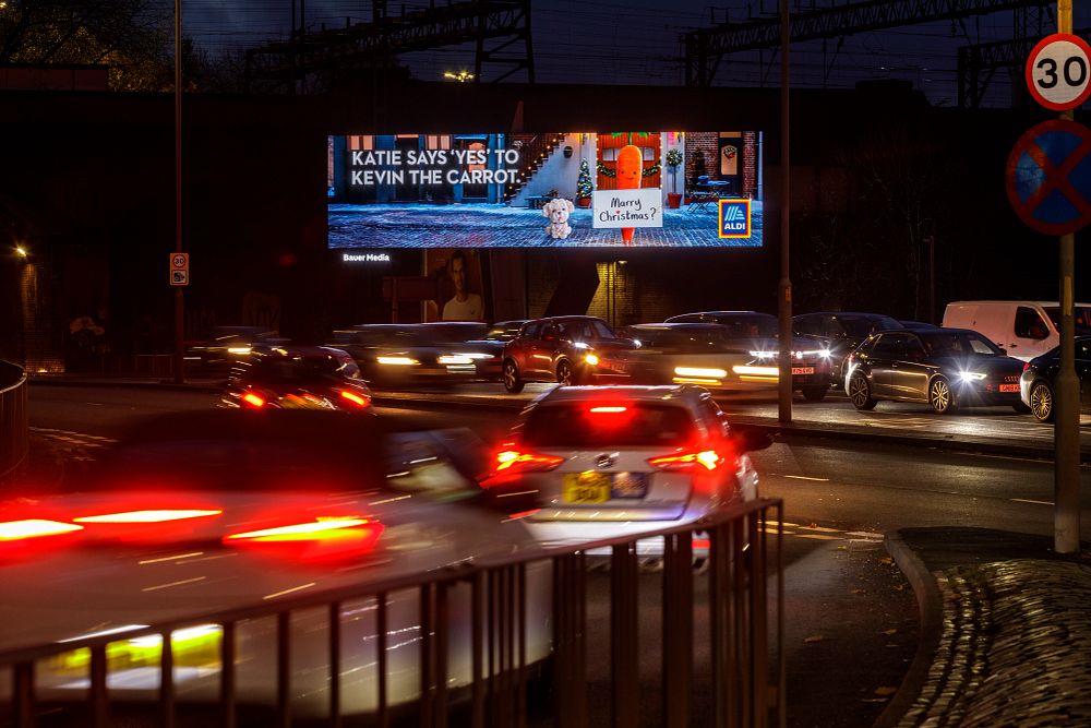 Nighttime traffic at a roundabout with a large illuminated billboard advertising Aldi, featuring a carrot and the text 'Katie says yes to Kevin the carrot' and 'Merry Christmas?'
