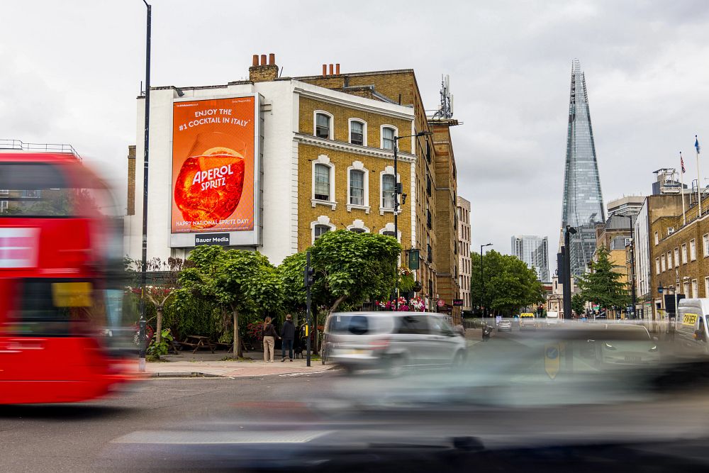 Busy London street scene with a moving red double-decker bus and cars, a brick building with a large Aperol Spritz billboard, and the Shard skyscraper in the background.