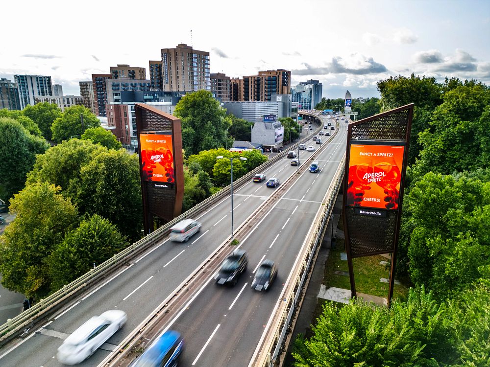 Elevated highway with fast-moving cars flanked by digital billboards advertising Aperol Spritz, surrounded by green trees and city buildings in the background.