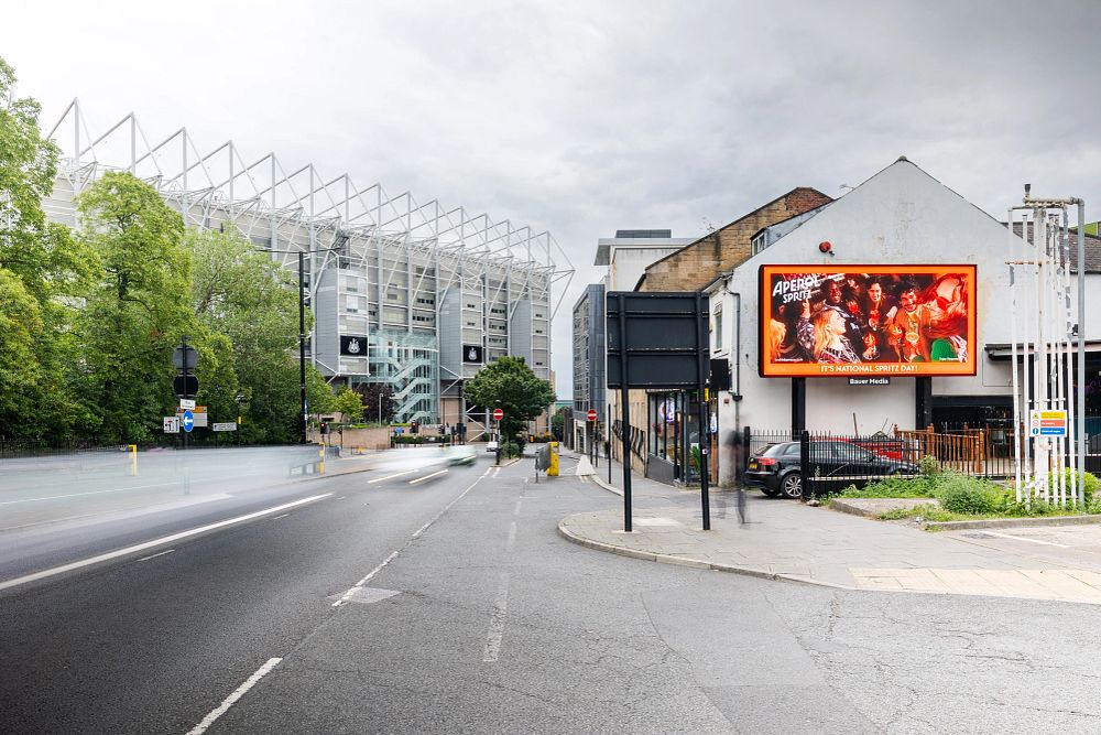 Street view with a large stadium on the left and a digital billboard on a building showing Aperol Spritz advertisement on the right.