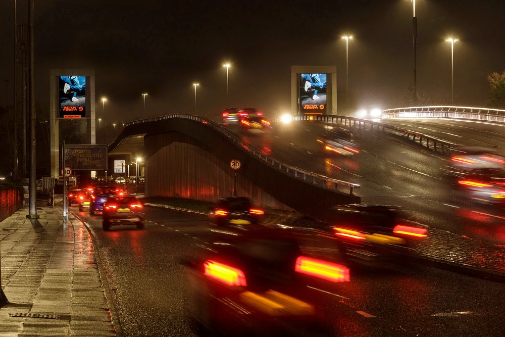 Blurred headlights and taillights of cars moving on a wet highway ramp at night under streetlights.