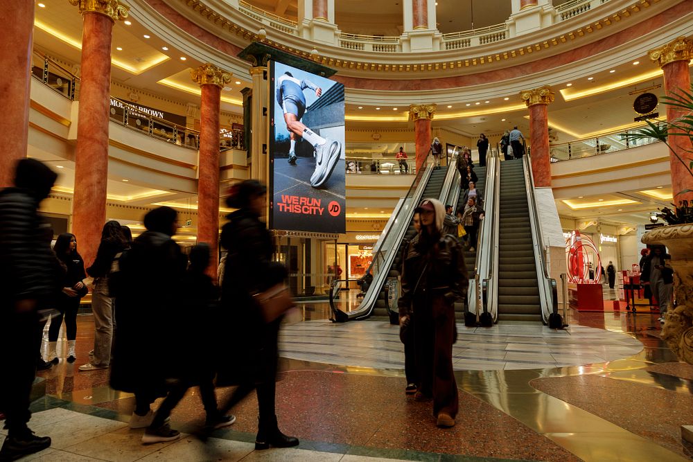 Busy shopping mall interior with people walking near escalators and a large digital billboard showing a runner's leg and shoe with 'We Run This City' advertisement.