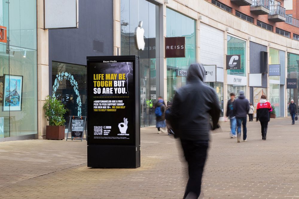 Urban shopping street with a digital billboard displaying a mental health support message for men, surrounded by pedestrians and retail stores.
