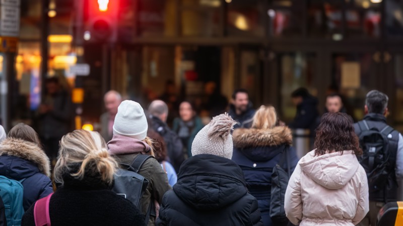 Crowd of people wearing winter coats and hats, standing and facing a red pedestrian traffic light in a city street.