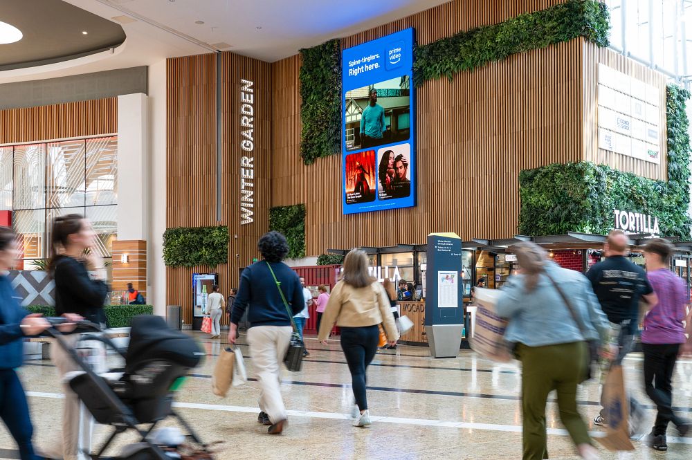 People walking inside a shopping mall near a wall with greenery and a Prime Video advertisement screen.