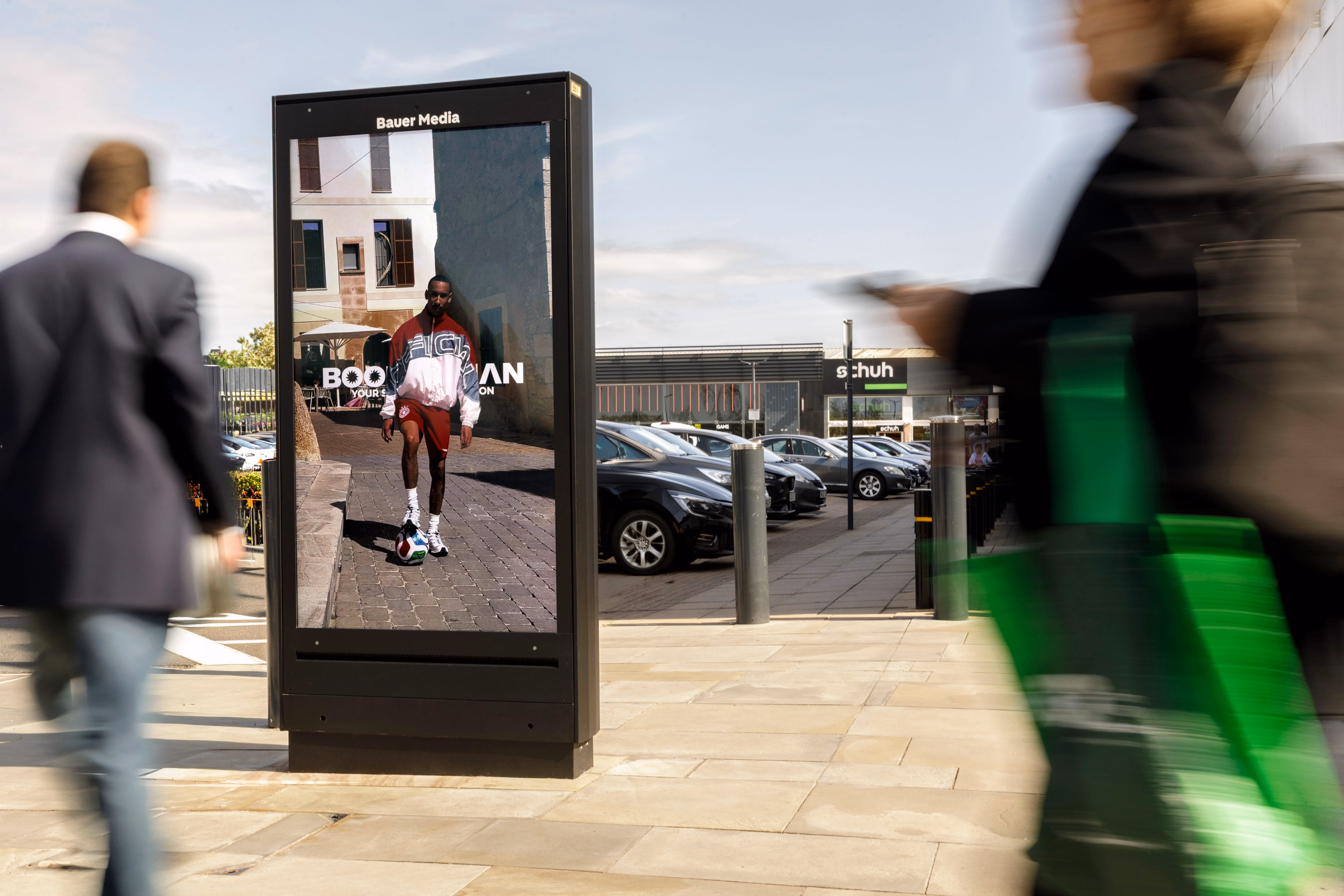 Digital billboard on sidewalk showing man in sportswear with a soccer ball, two blurred pedestrians passing.