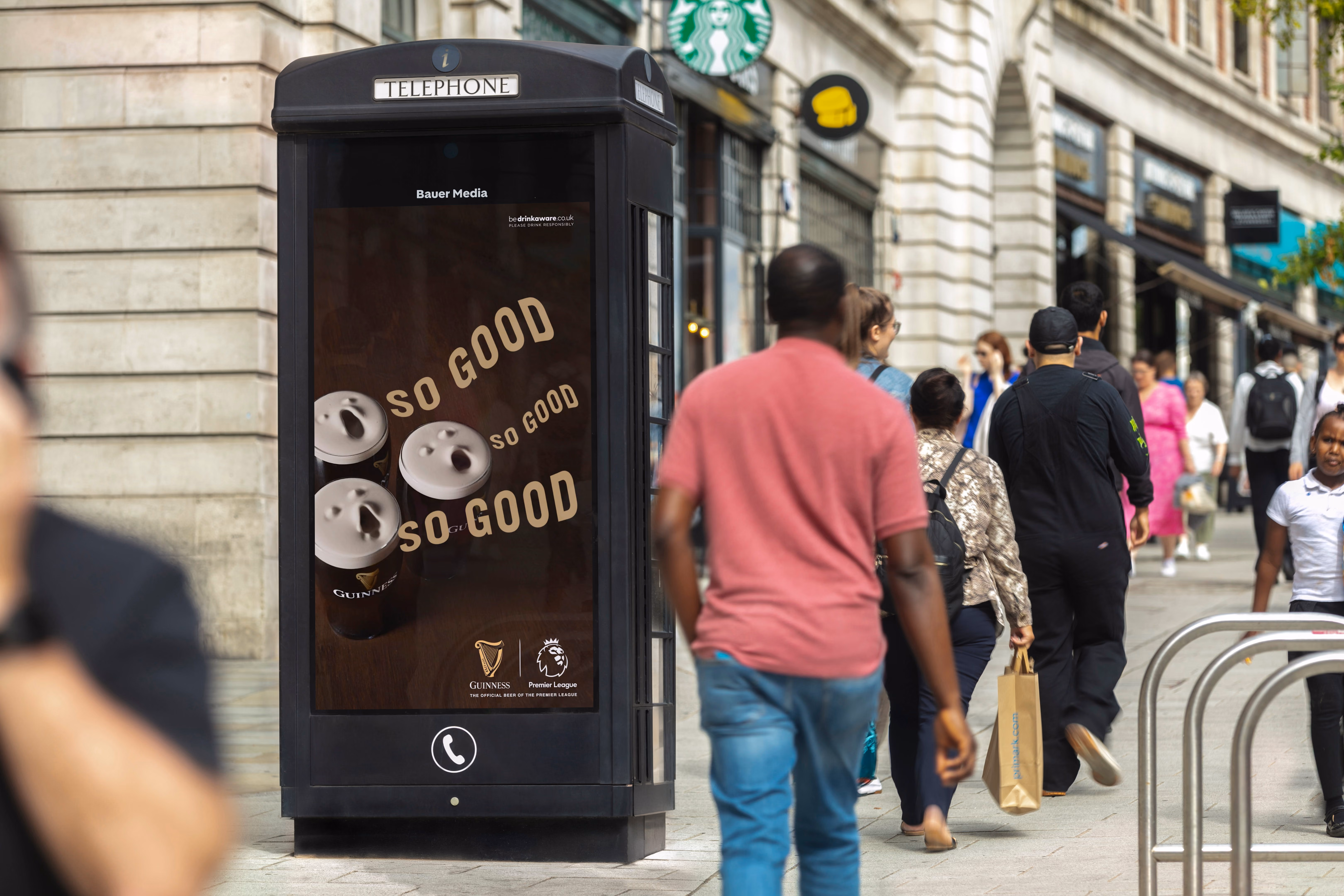 Black telephone booth on city street with Guinness ad showing three pints and words 'SO GOOD'.