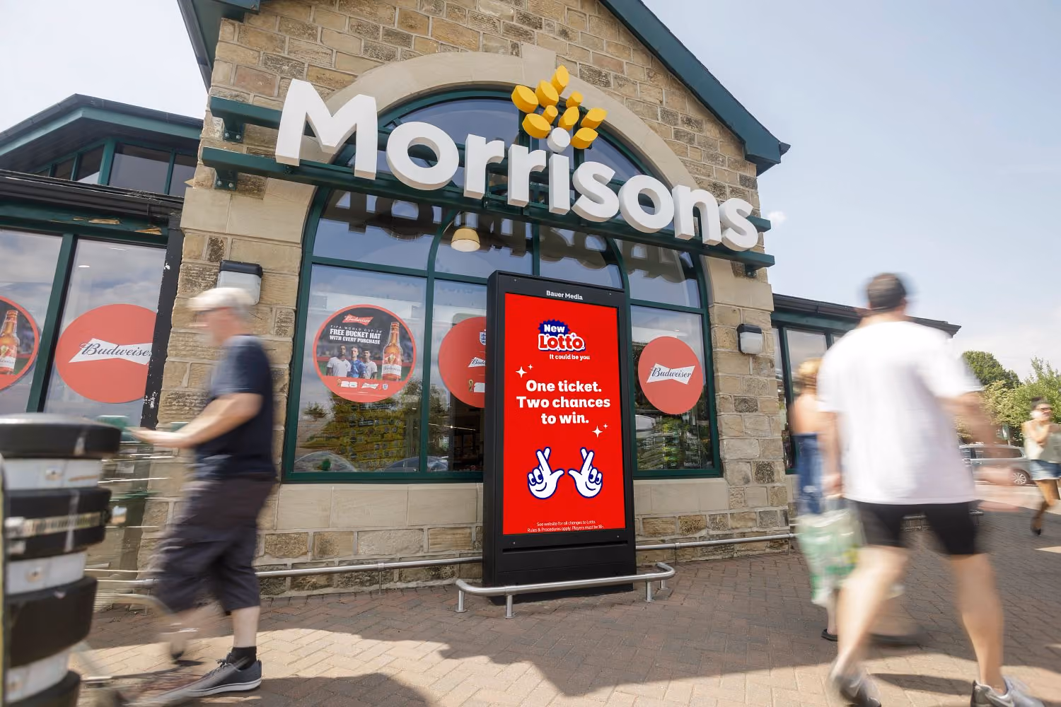 Exterior of a Morrisons supermarket with a red digital display advertising New Lotto, stating 'One ticket. Two chances to win.' People are walking by with shopping trolleys and bags.
