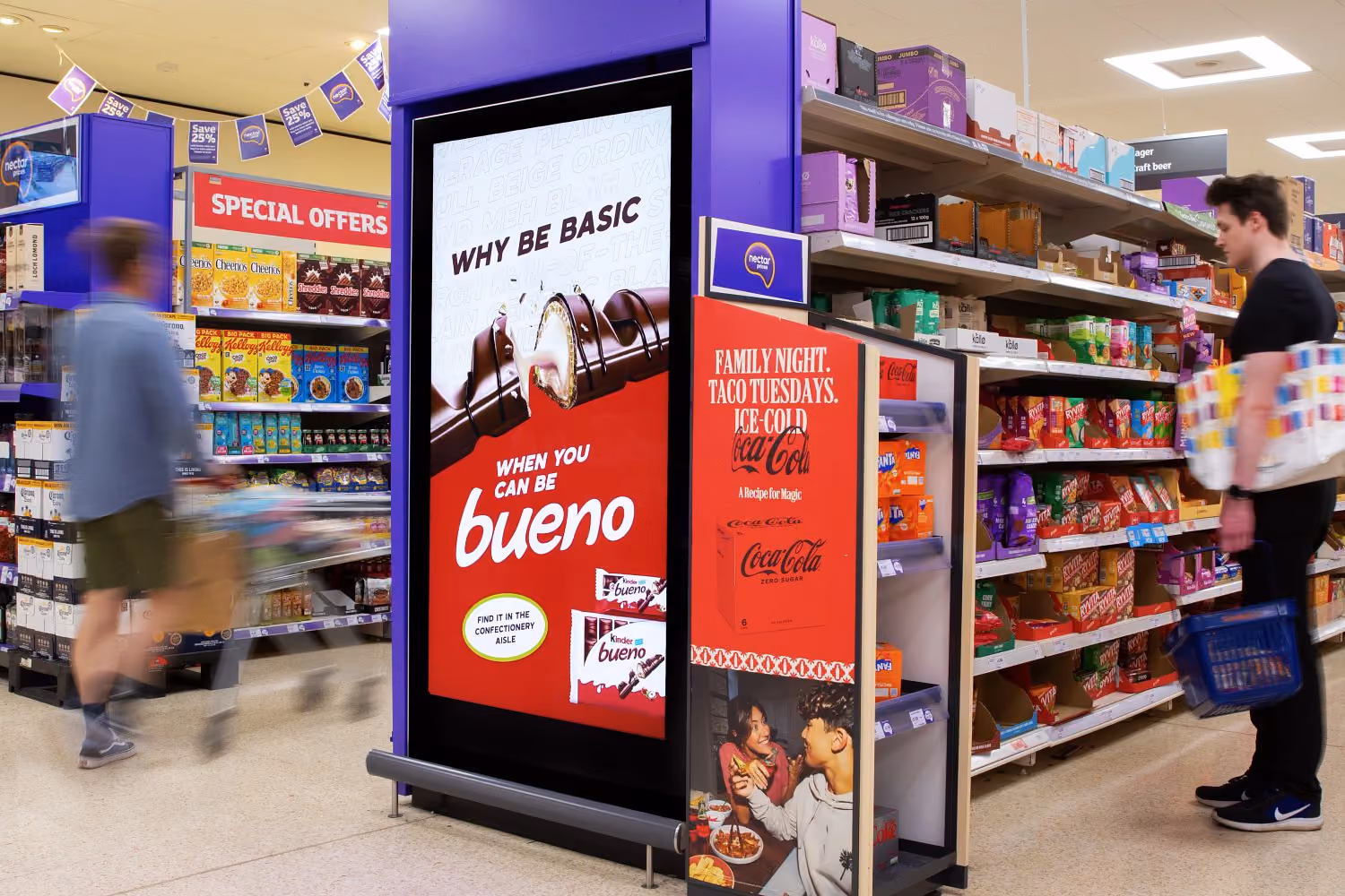 Supermarket aisle with shoppers; a digital display prominently features a Kinder Bueno chocolate bar ad with the slogan 'Why be basic when you can be bueno.'