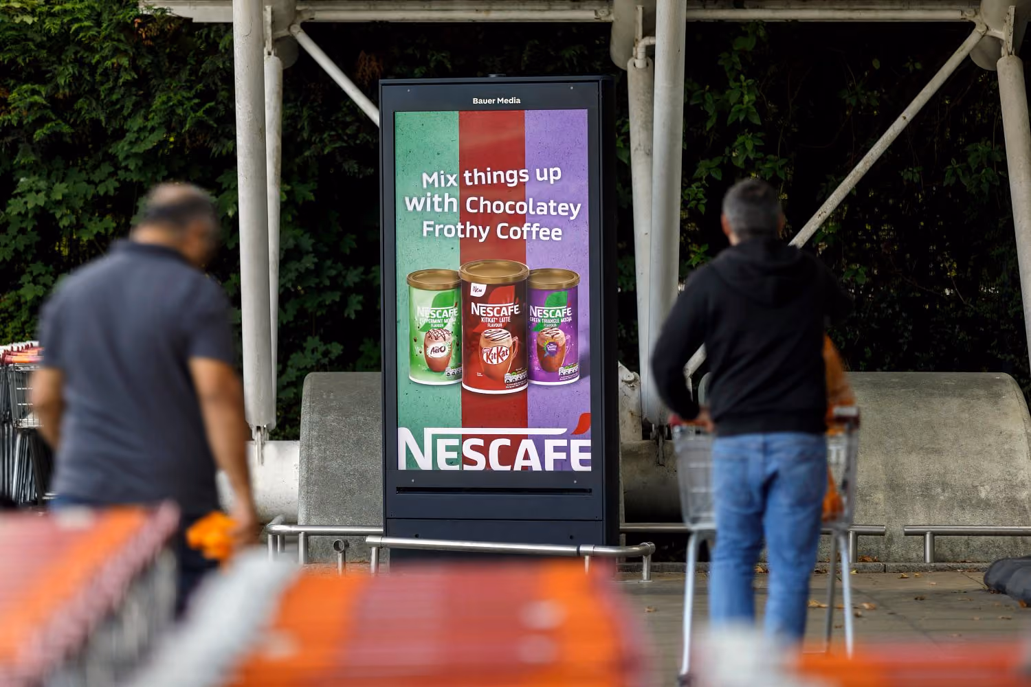 Outdoor digital advertisement displaying three cans of Nescafé coffee with the text 'Mix things up with Chocolatey Frothy Coffee' and the Nescafé logo at the bottom, with two people pushing shopping carts passing by.