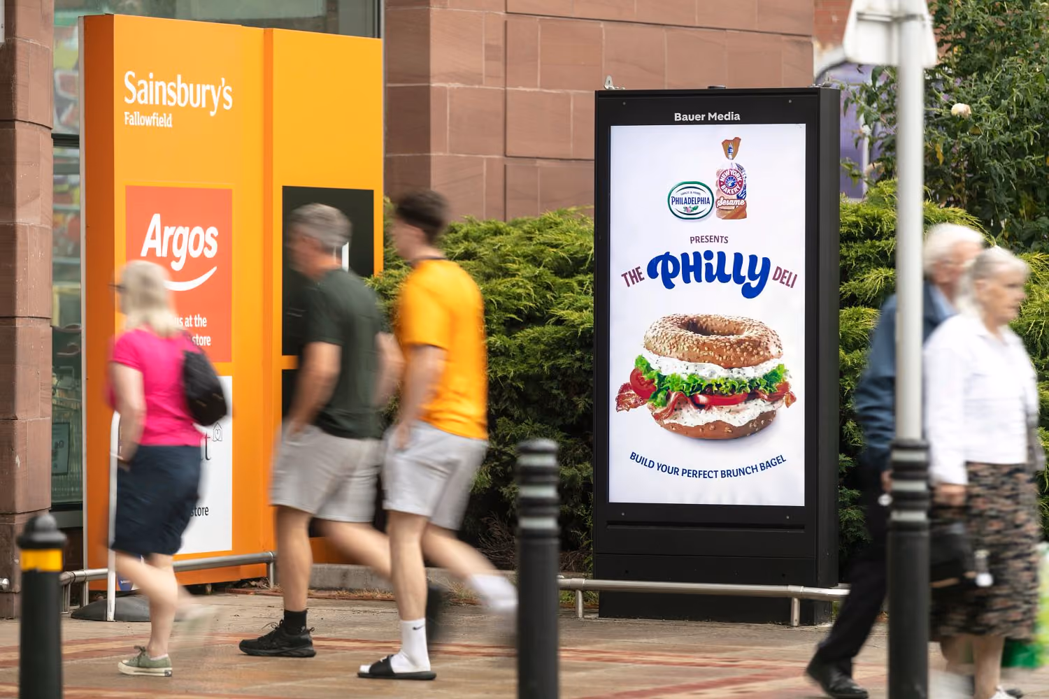Outdoor digital advertisement for The Philly Deli bagel showing a bagel sandwich with lettuce, tomato, and cream cheese, promoting building your perfect brunch bagel, near a Sainsbury's and Argos store entrance with blurred pedestrians walking by.