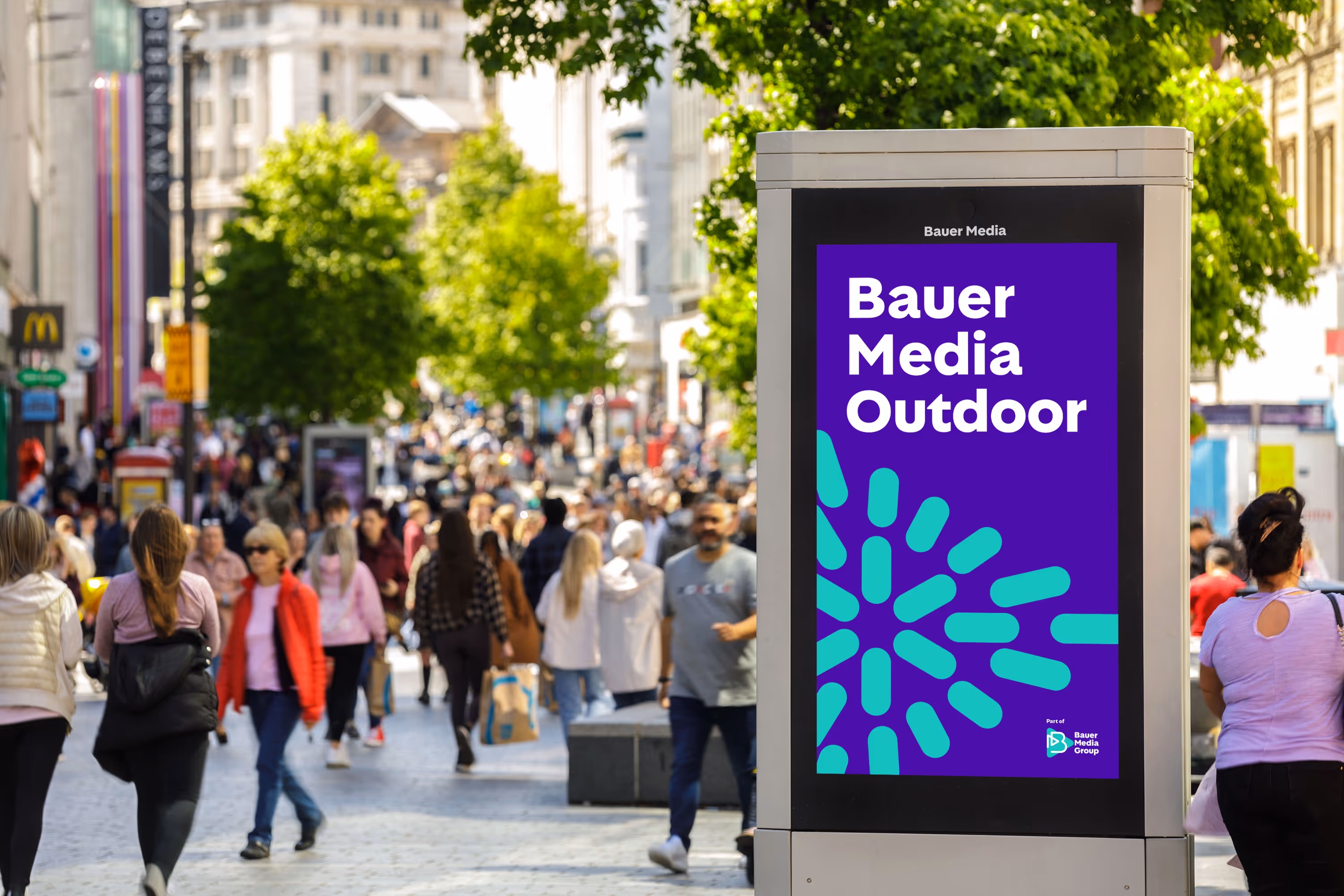 Bauer Media Outdoor advert on an Adshel, on busy sidewalk as pass by