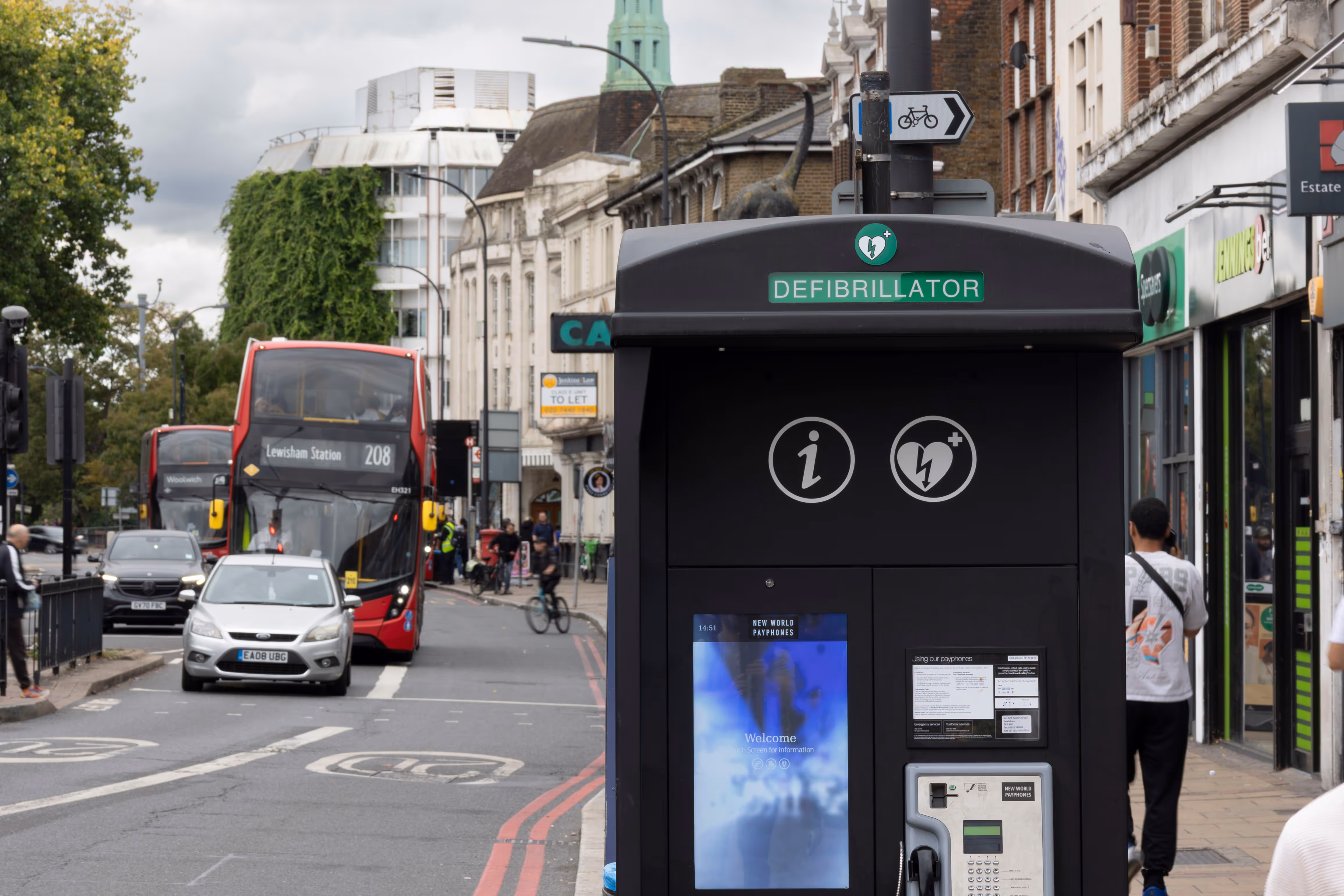 A communication kiosk on a busy sidewalk as pedestrians and vehicles pass by during the day.
