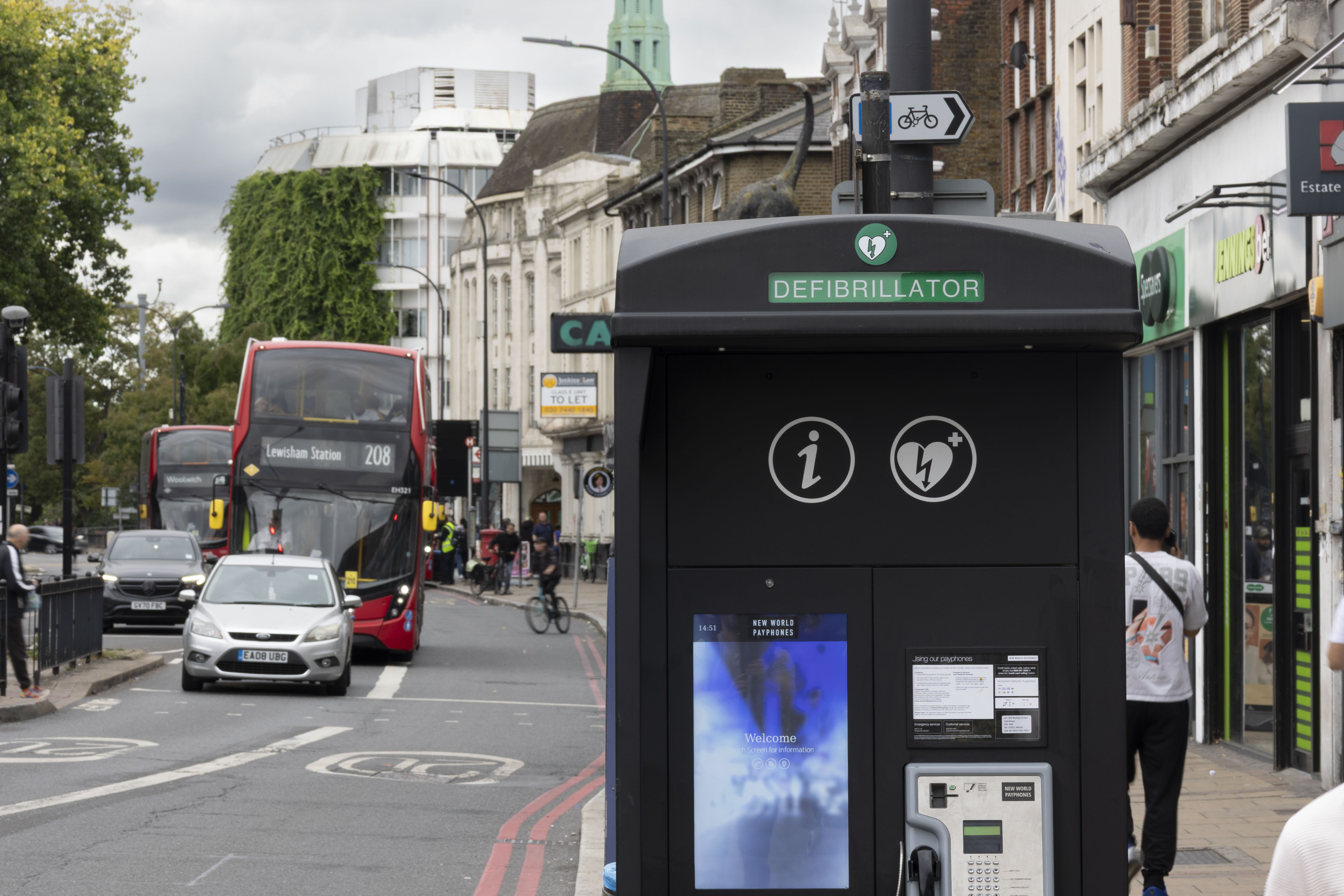 A communication kiosk on a busy sidewalk as pedestrians and vehicles pass by during the day.