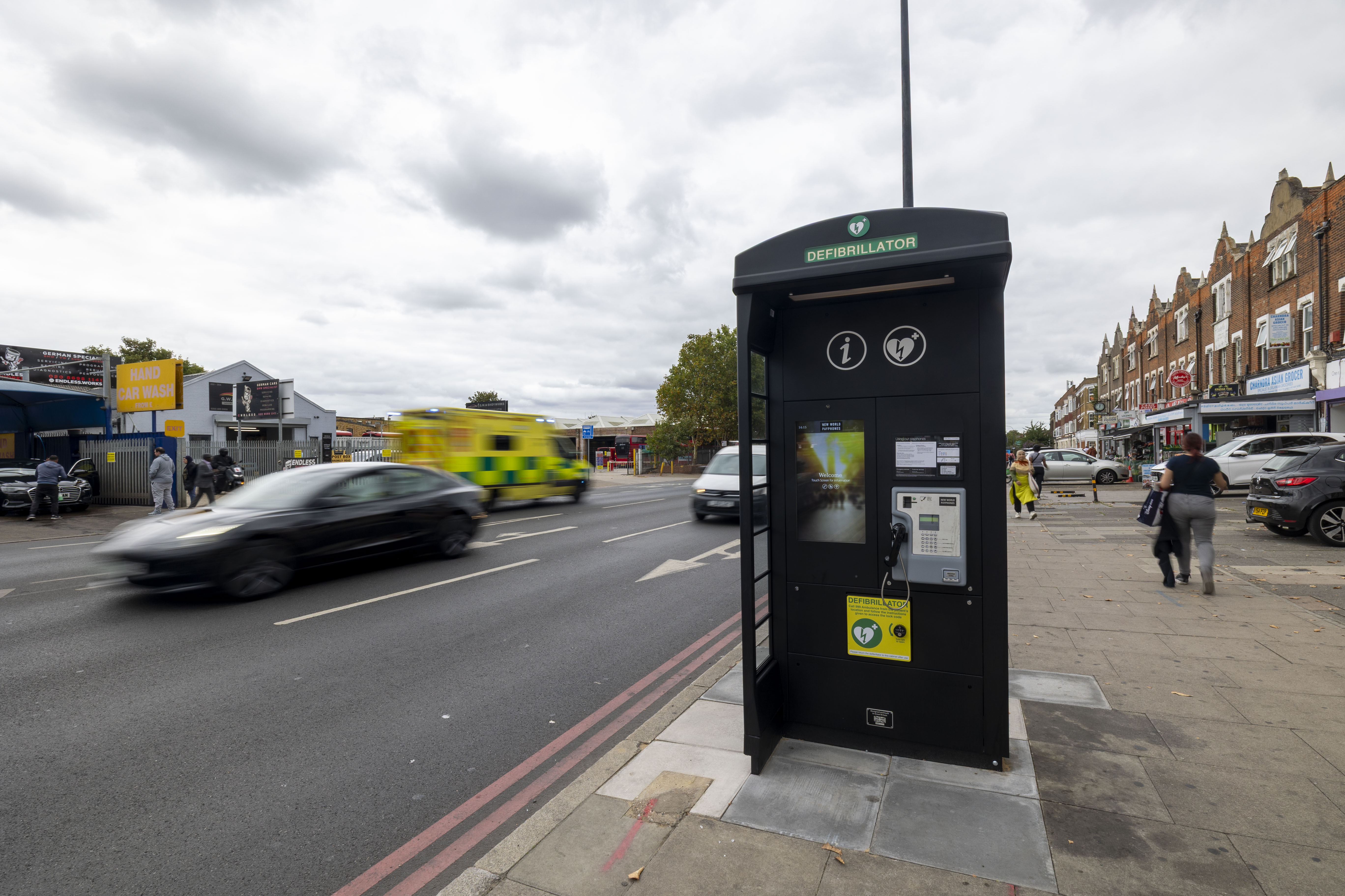 A communication kiosk, featuring a defibrillator, on a busy sidewalk as pedestrians and vehicles pass by during the day.