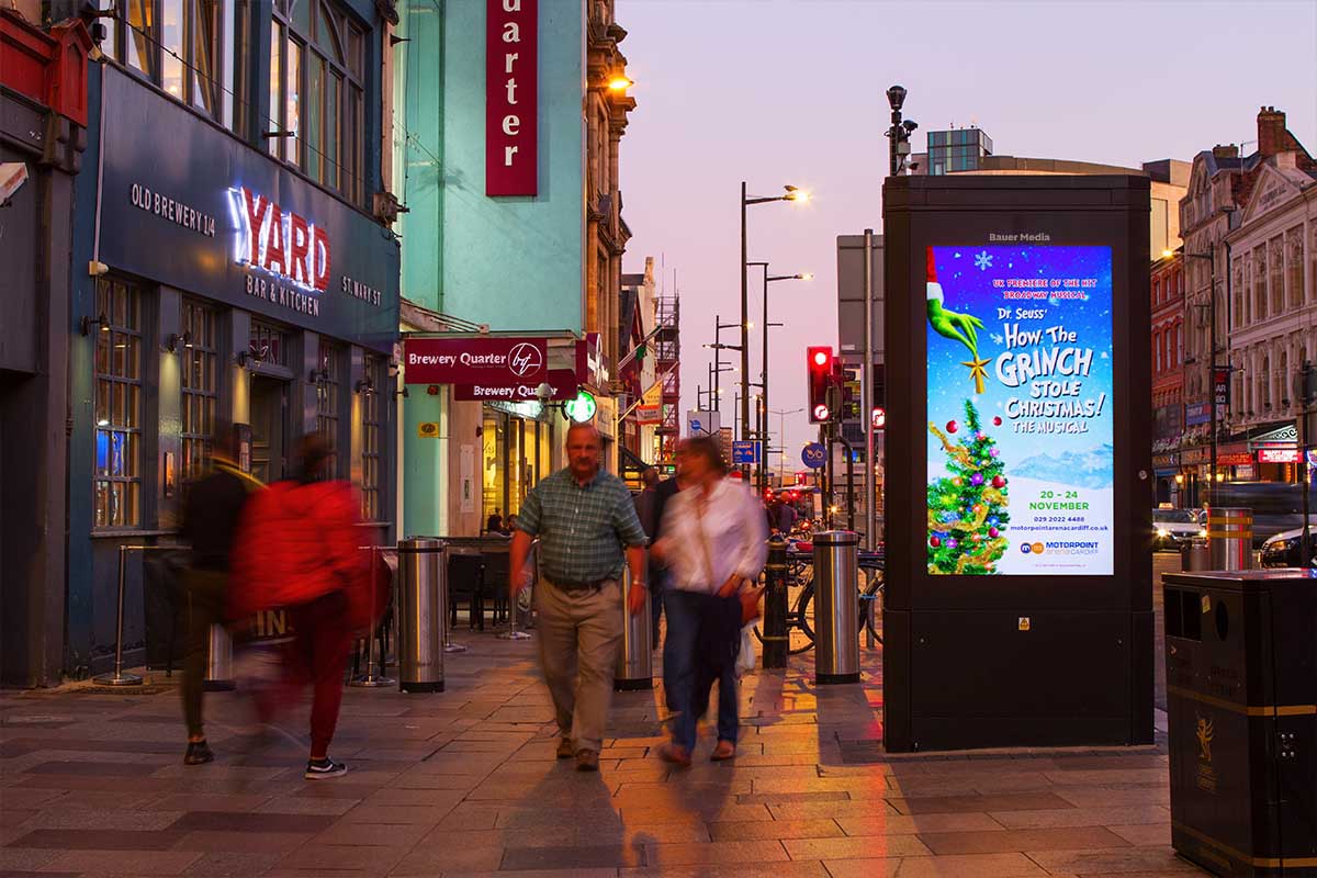 Pedestrians walk past a Bauer Media digital screen, on a busy sidewalk, at dusk