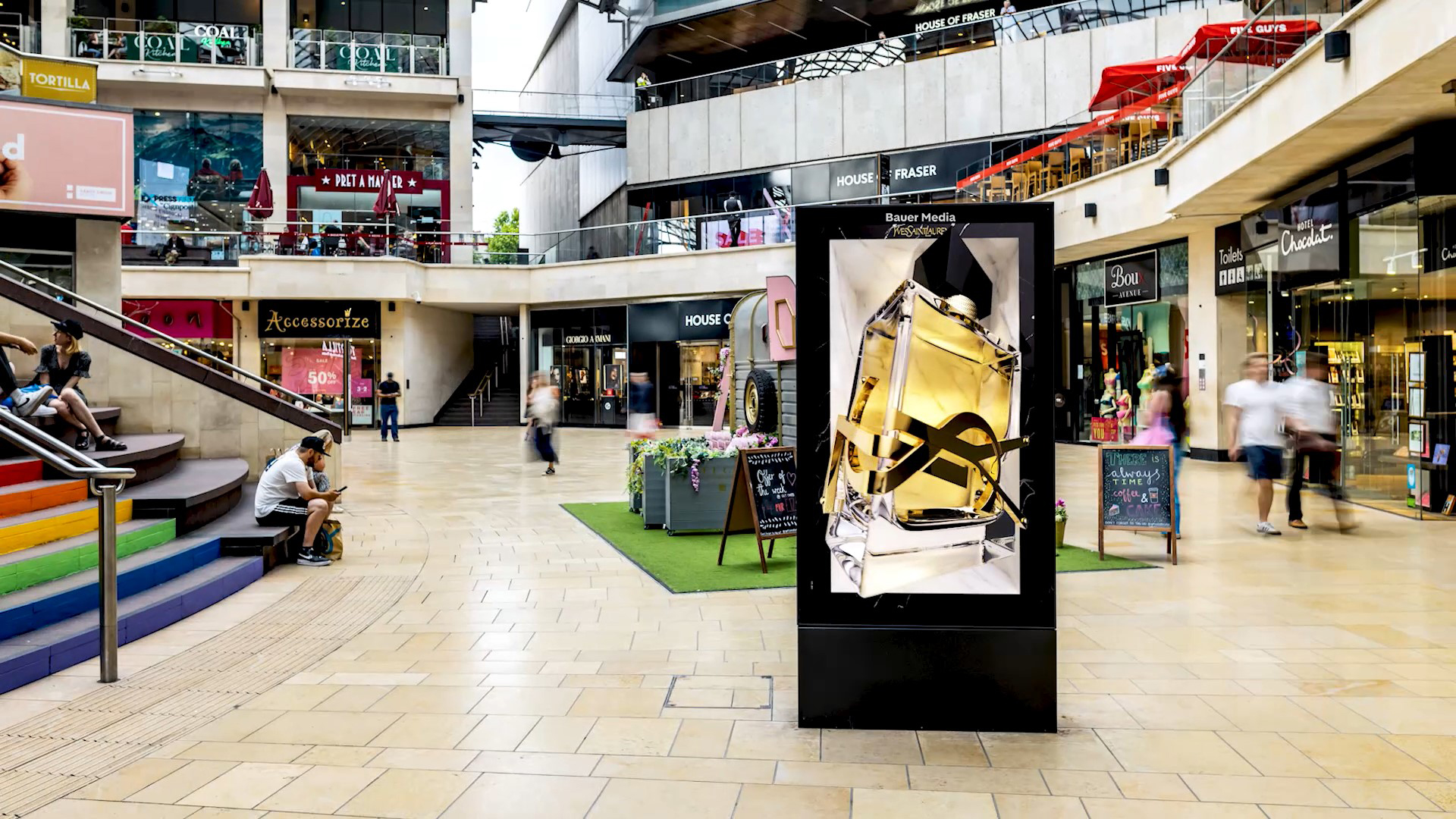 A YSL 3D build features on a digital screen in a shopping centre, during the day, as shoppers pass by