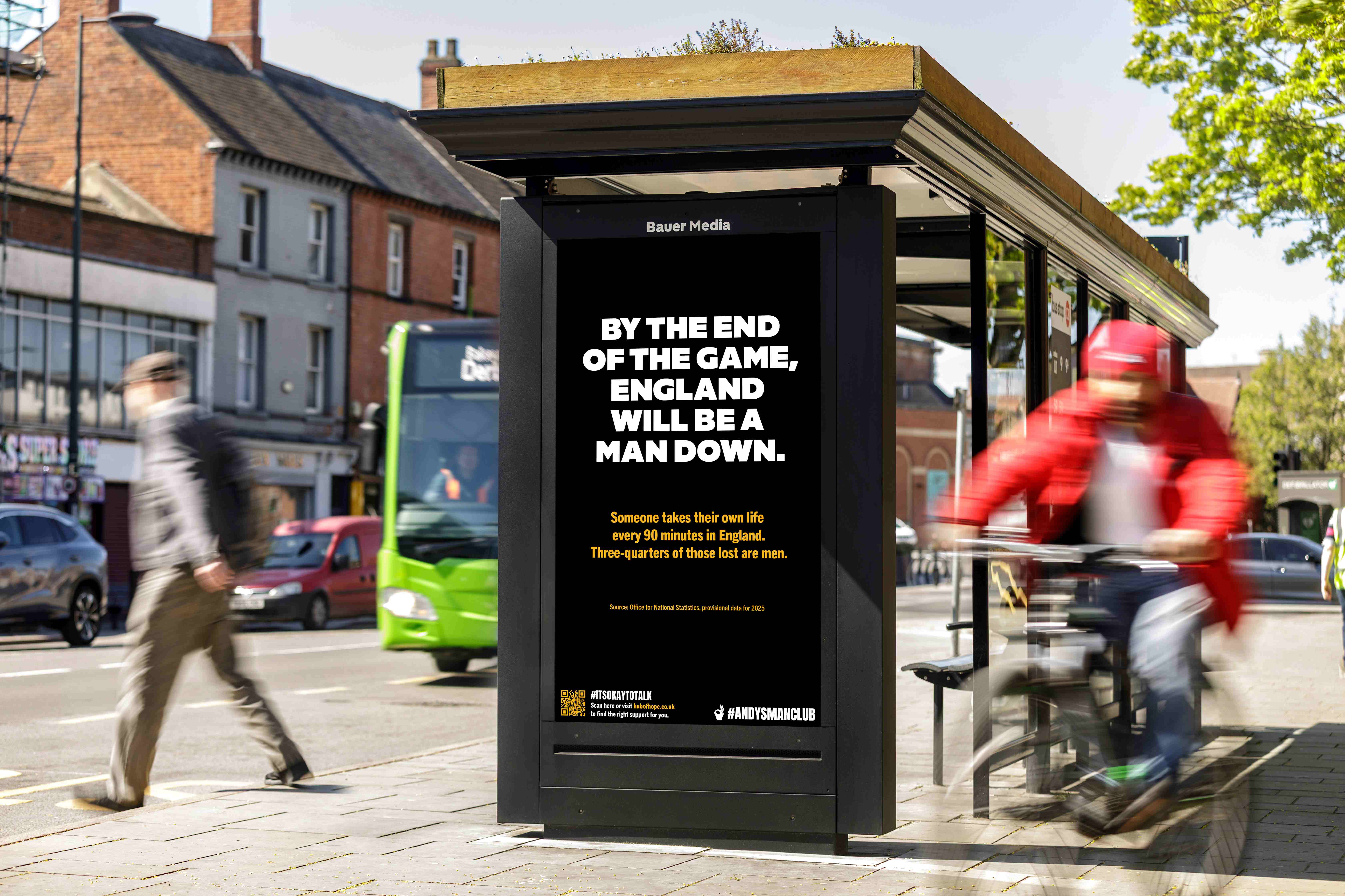 Bauer Media Outdoor bus shelter featuring an ANDYSMANCLUB advert, on a busy sidewalk as pedestrians and vehicles pass by.