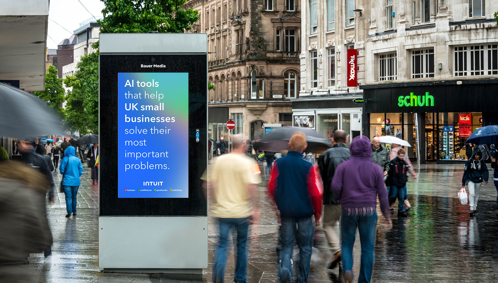 Bauer Media Outdoor digital kiosk featuring an adshel, on a rainy city shopping street as pedestrians with umbrellas pass by.