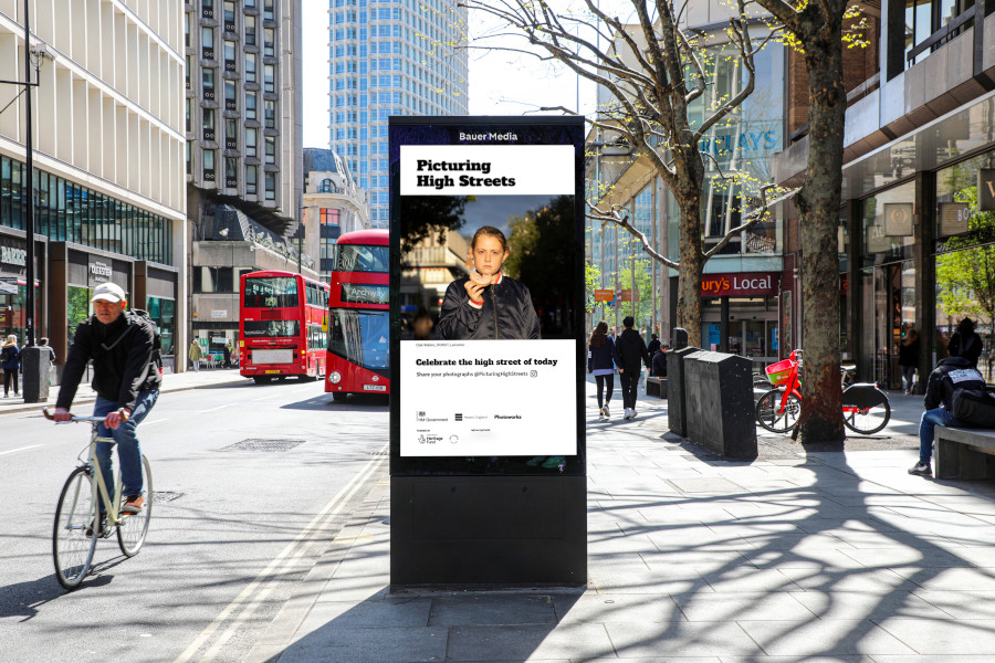Bauer Media Outdoor Adshel featuring a Picturing High Streets advert, on a sunny city street as cyclists, pedestrians and red buses pass by.