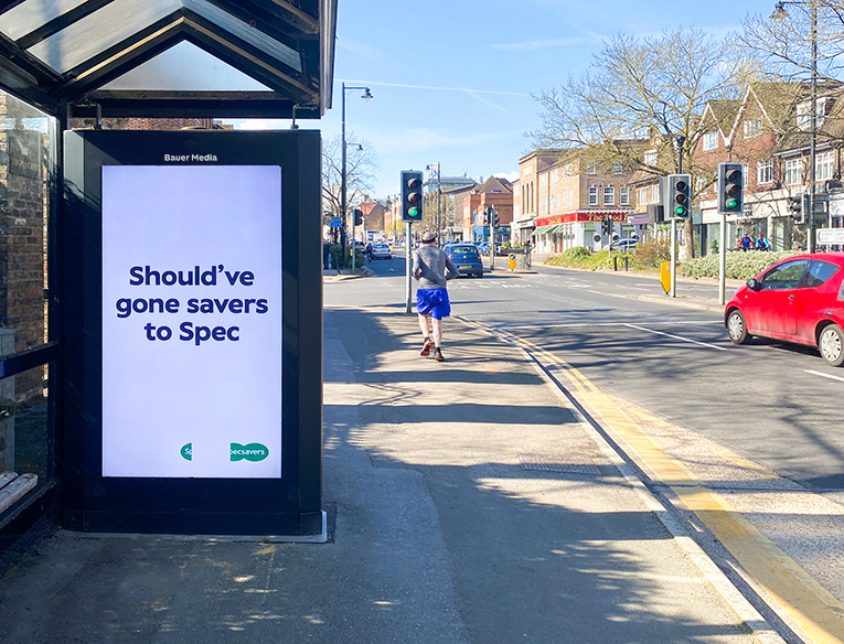 Bauer Media digital billboard with a Specsavers advert on a sunny high street, beside a road junction and passing traffic.