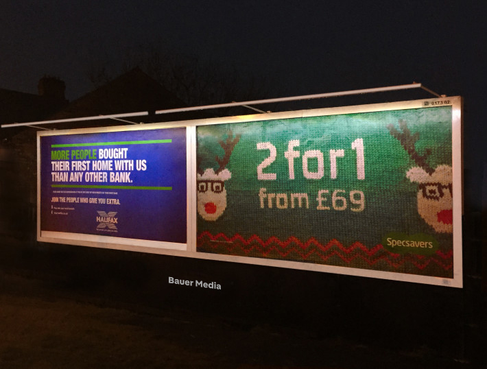 A lit roadside billboard at night displays two adverts: one for Halifax about first-time buyers and one for Specsavers promoting “2 for 1” glasses with a festive reindeer knit design.