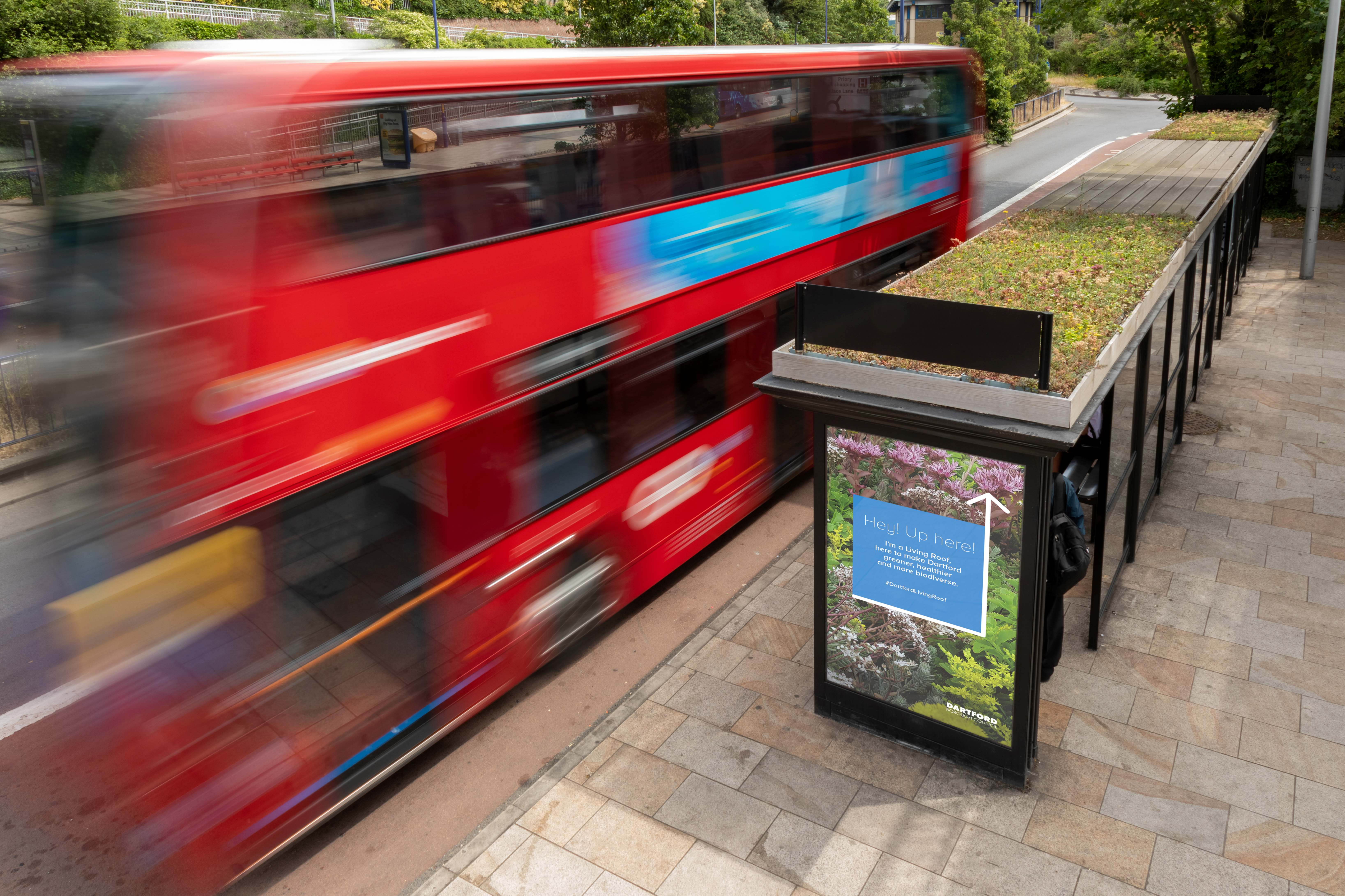 A red double-decker bus passes a bus stop with a digital advertising display on a city street.
