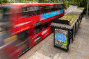 A red double-decker bus passes a bus stop with a digital advertising display on a city street.