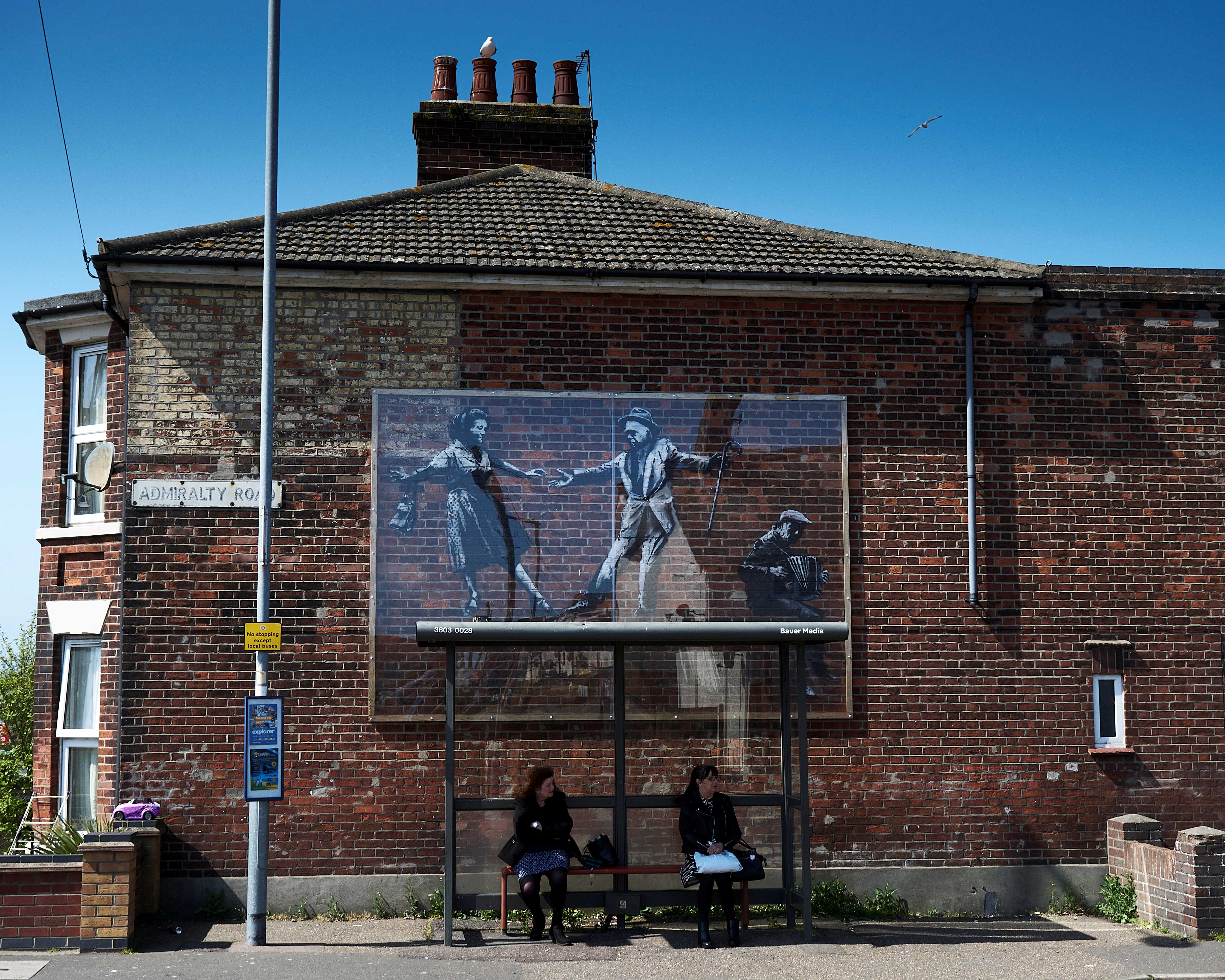 A mural by Banksy on a wall behind two people waiting at a bus stop