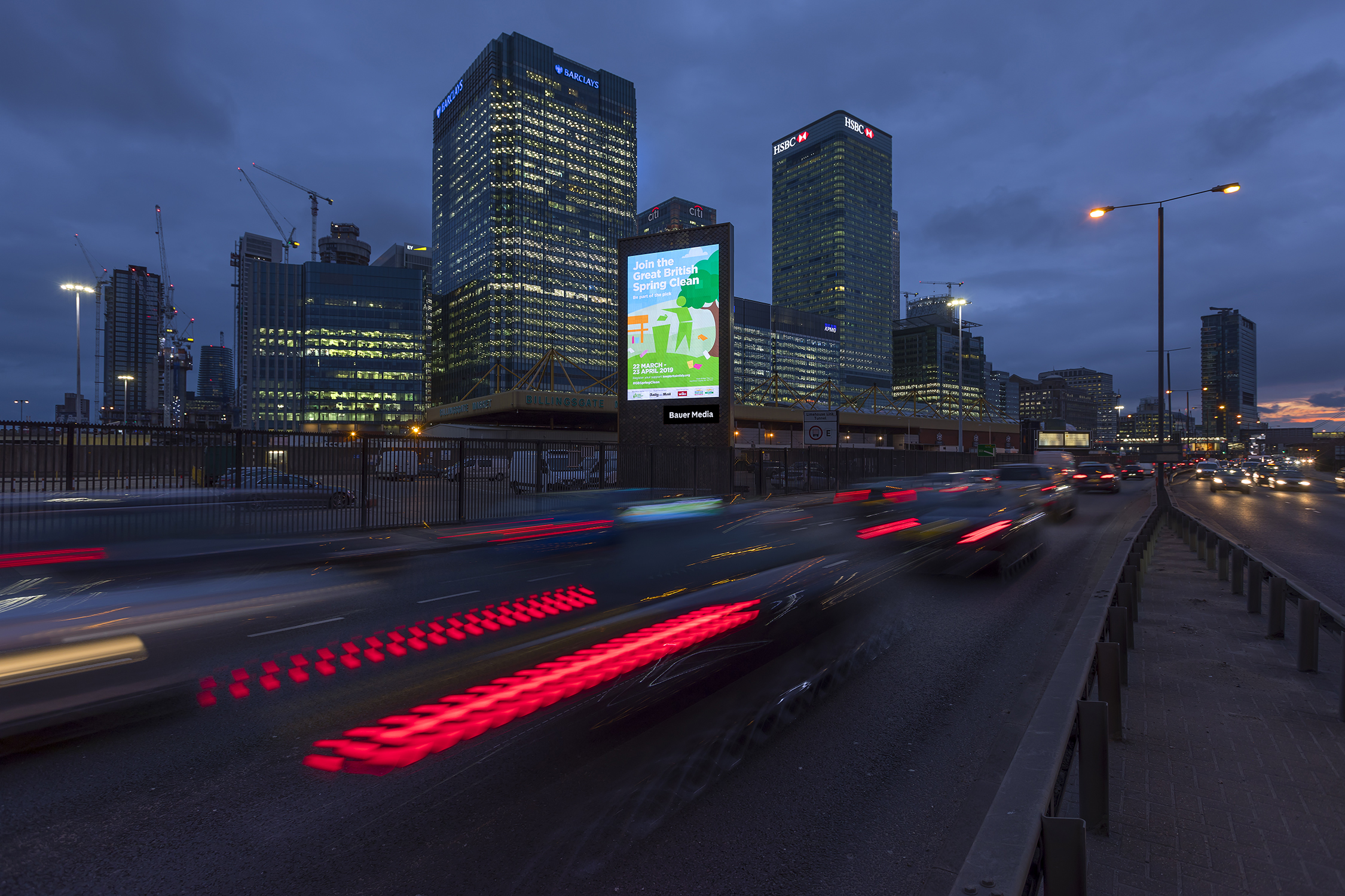 A illuminated 'Great British Spring Clean' billboard on a busy dual carriageway at dusk, with light trails from passing traffic and the Canary Wharf skyline behind.