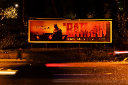 A illuminated billboard displaying a dark, red-toned advertisement photographed at night, with light trails from passing traffic visible in the foreground.
