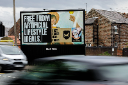 A roadside Bauer Media billboard advertising Beavertown’s “Lost Lager” with the headline “Free from artificial lifestyle ideals,” shown beside brick buildings with blurred traffic passing in the foreground.