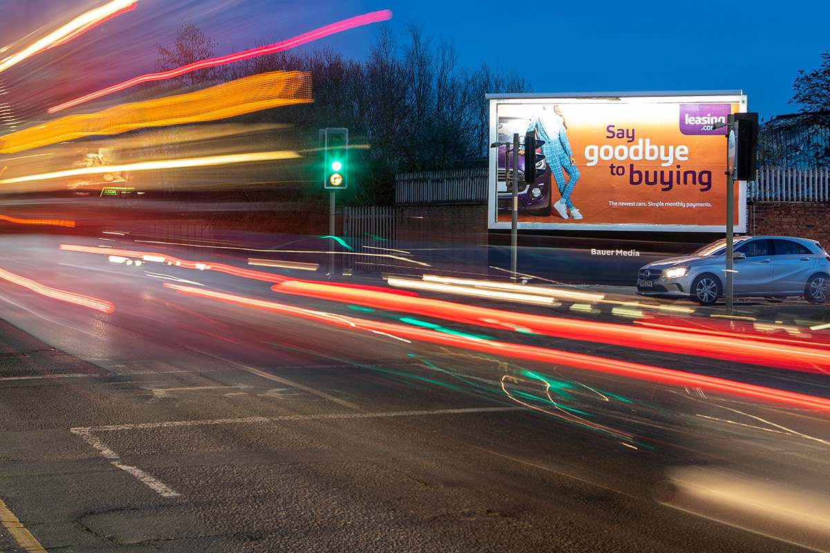 A nighttime roadside billboard advertising Legends of Gaming beside a busy road, with long-exposure light trails from passing traffic stretching across the scene.
