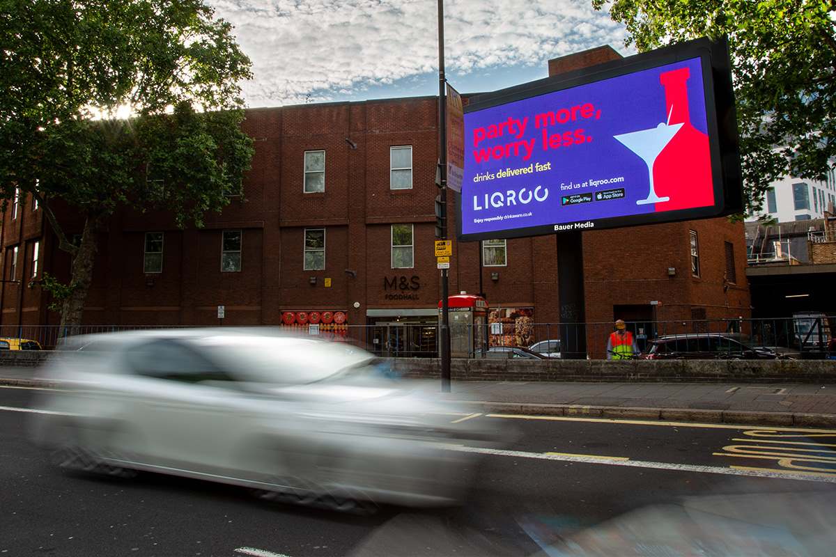 A digital roadside billboard in an urban street setting displaying a bright red and blue entertainment advert, with a blurred white car passing in the foreground.