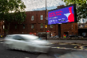 A digital roadside billboard in an urban street setting displaying a bright red and blue entertainment advert, with a blurred white car passing in the foreground.