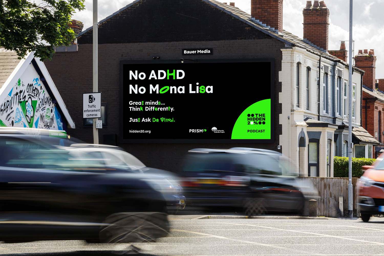 A roadside billboard on the side of a building displaying a dark green advert for KISS, with blurred cars passing in the foreground on a residential street.