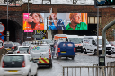 A large digital billboard in Manchester displays a colourful Very advert reading “Be very you,” positioned above a busy road with traffic passing in front and a railway bridge behind it.