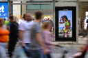 Blurred commuters walking past a digital advertising screen in a busy indoor station or shopping area.