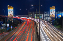 Long-exposure night view of a busy motorway, with red and white light trails from traffic passing between illuminated roadside signs.