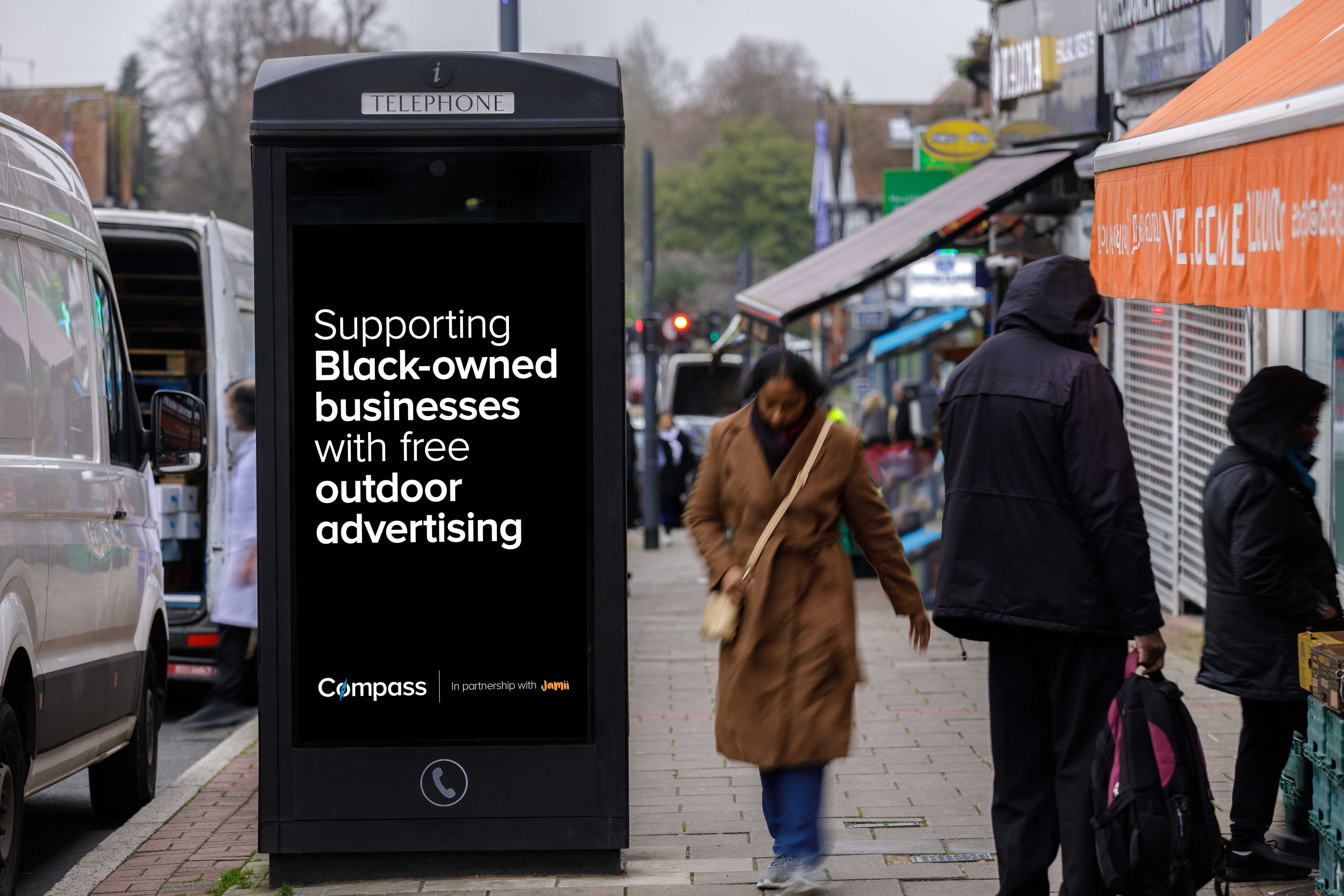 A digital advertising screen in a black telephone kiosk on a busy high street displays a message supporting Black-owned businesses with free outdoor advertising, as pedestrians pass by.