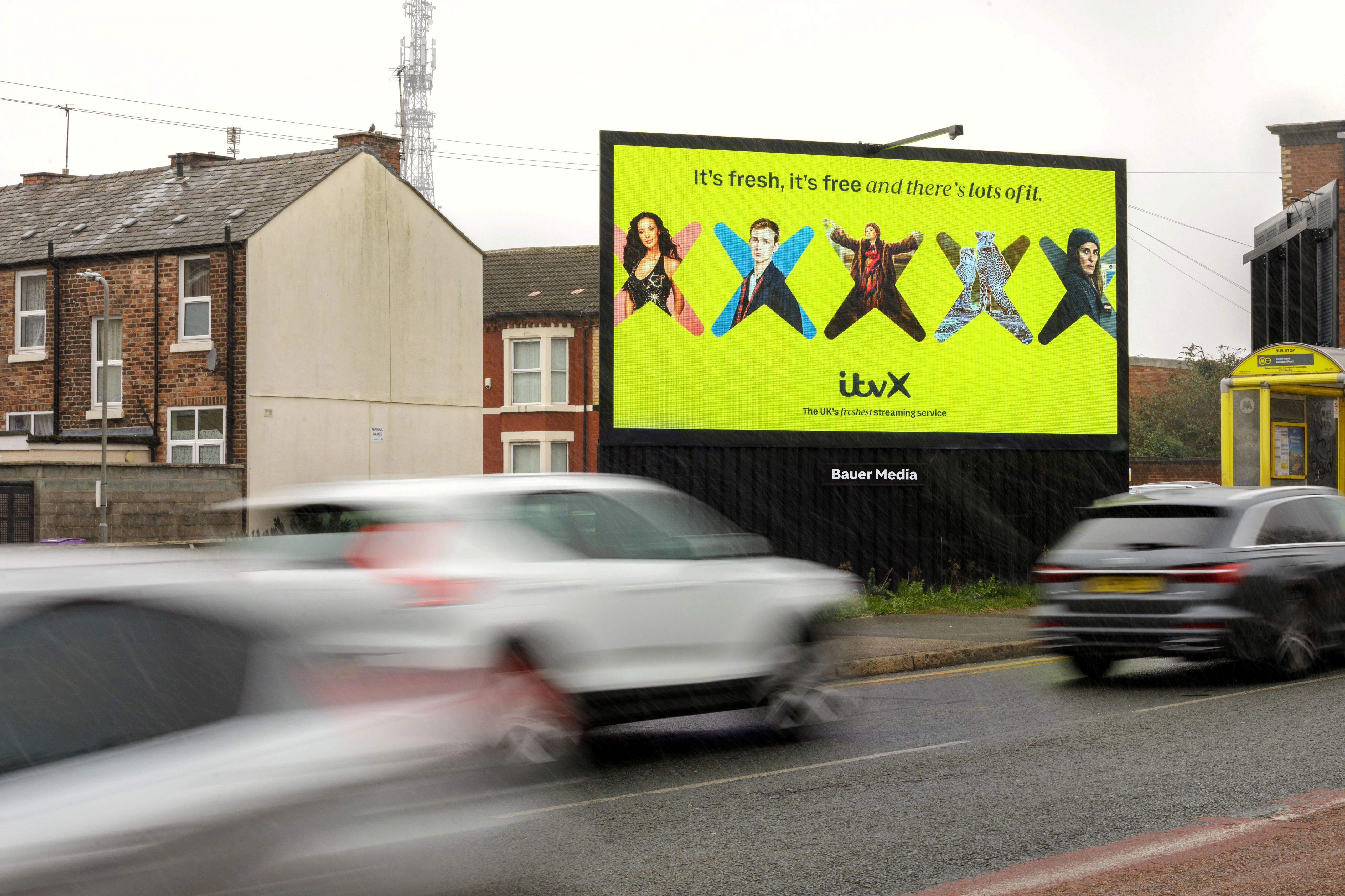 A bright yellow ITVX billboard on a roadside, advertising “It’s fresh, it’s free and there’s lots of it,” with blurred traffic passing in front on a rainy urban street.
