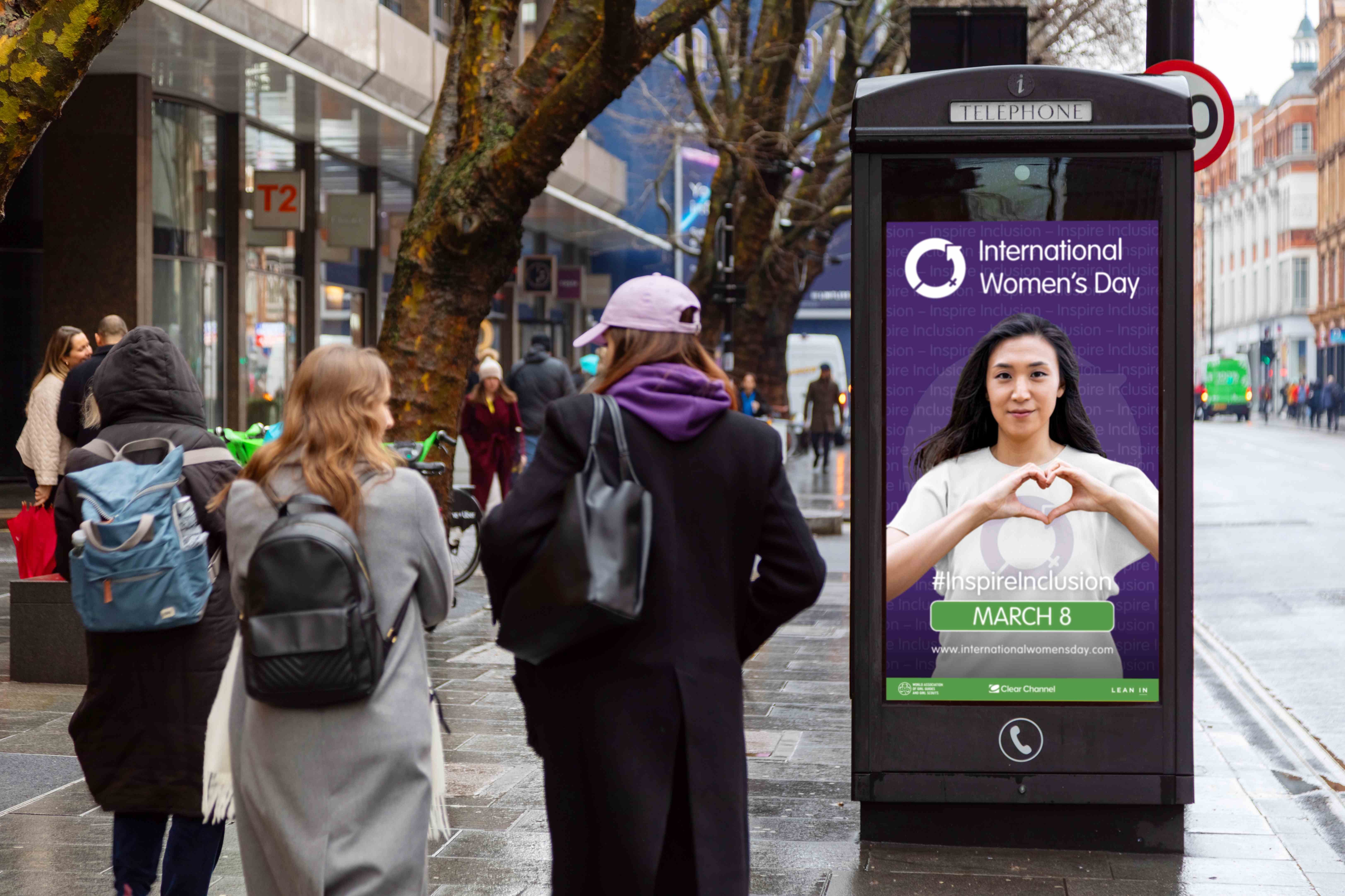 Pedestrians walk along a city pavement past a digital phone box advert for International Women’s Day, showing a person making a heart shape with their hands and the message “Inspire Inclusion.”