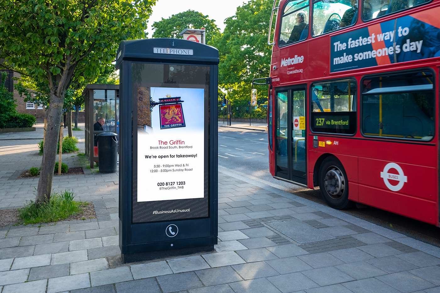 A digital phone box advert for The Griffin pub stands beside a bus stop, with a red double-decker bus passing on a tree-lined street.
